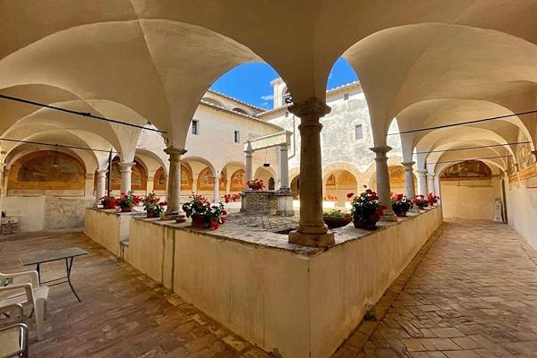 Cloister of the Church of San Francesco in Lugnano in Teverina with arched columns, a central well, and blooming geranium plants.