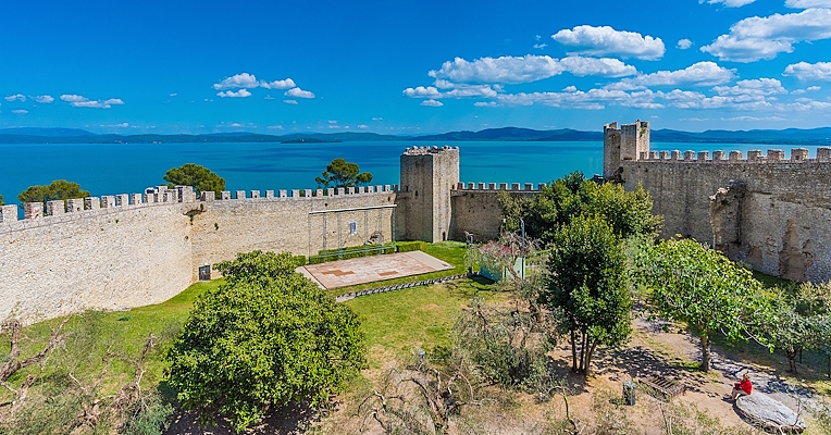 Vue d'ensemble de la Rocca del Leone, avec ses imposants remparts entourant des tours médiévales et un amphithéâtre en plein air à l'intérieur. En arrière-plan, le lac Trasimène et ses îles créent un paysage pittoresque.