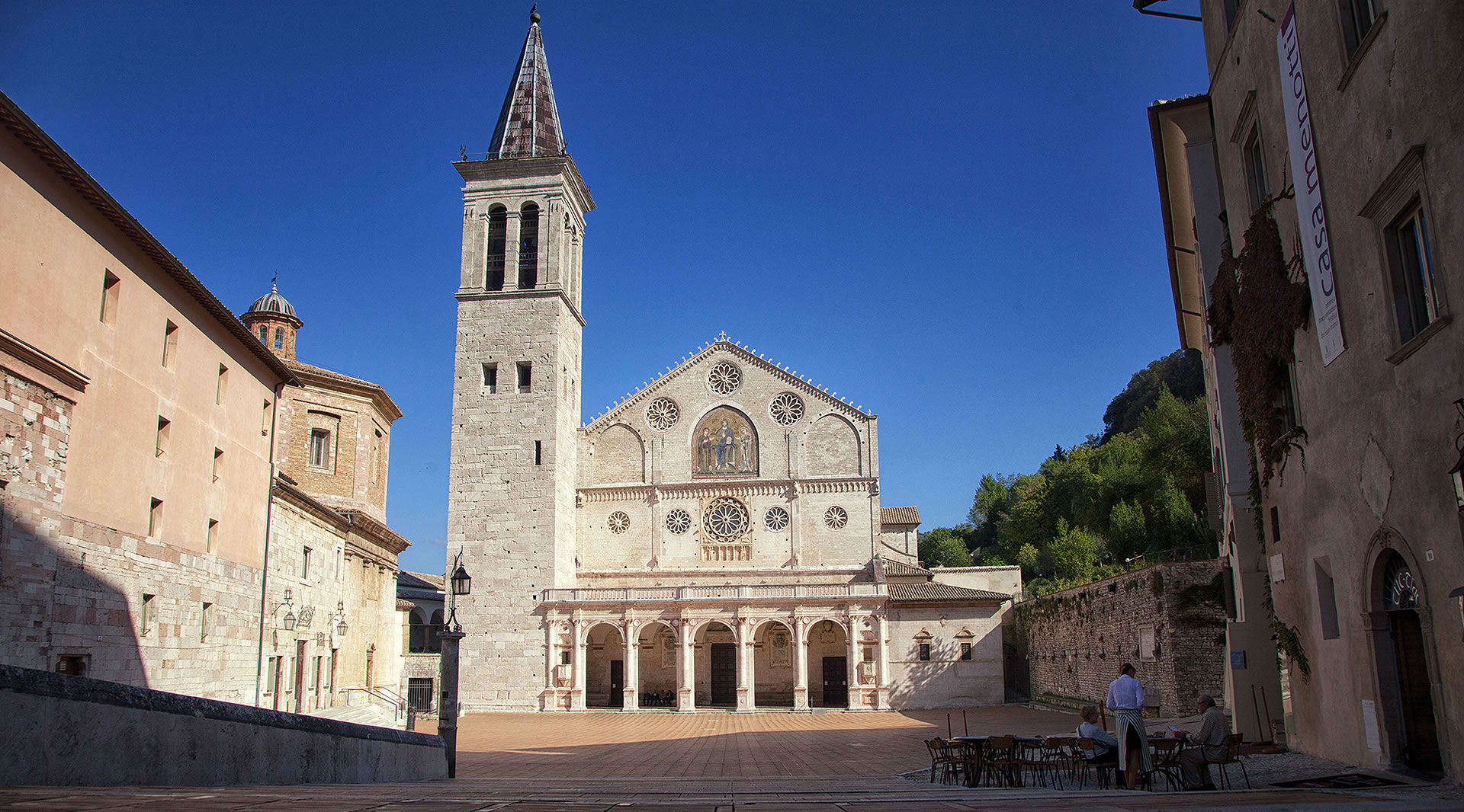 Spoleto Cathedral in white and pink stone, with a Romanesque façade, rose windows, and a portico, overlooking a sunny square.