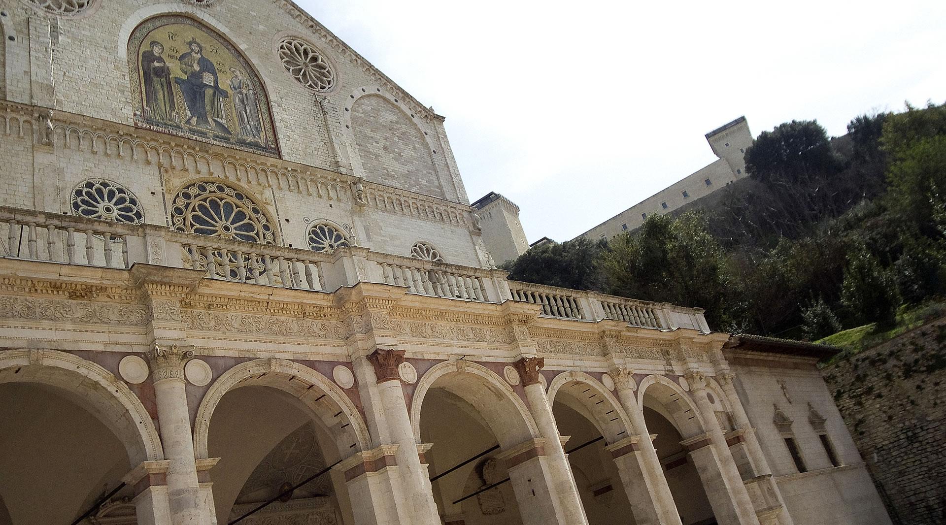 Facciata del Duomo di Spoleto con portico ad archi, rosoni, mosaico dorato e la Rocca Albornoziana sullo sfondo.