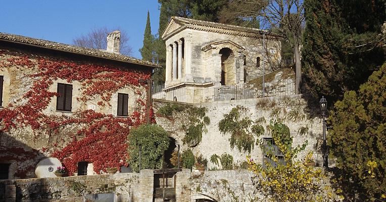 The Temple of Clitunno standing above and behind an ivy-covered building next to a canal