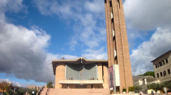  Modern facade of the Collevalenza Sanctuary with a tall brick bell tower, a blue sky, and clouds in the background. 