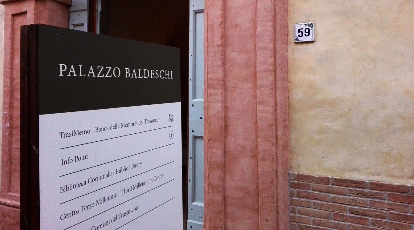 Vista del portone d'ingresso di Palazzo Baldeschi, con un cartellone informativo in primo piano che fornisce dettagli sul palazzo e le sue attività.