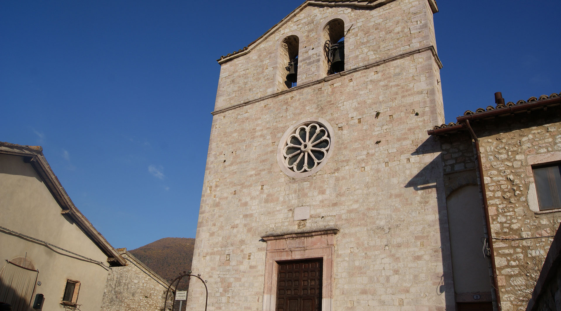 Stone façade of the Church of San Giovanni Battista in Vallo di Nera, with a bell gable, central rose window and clear sky.