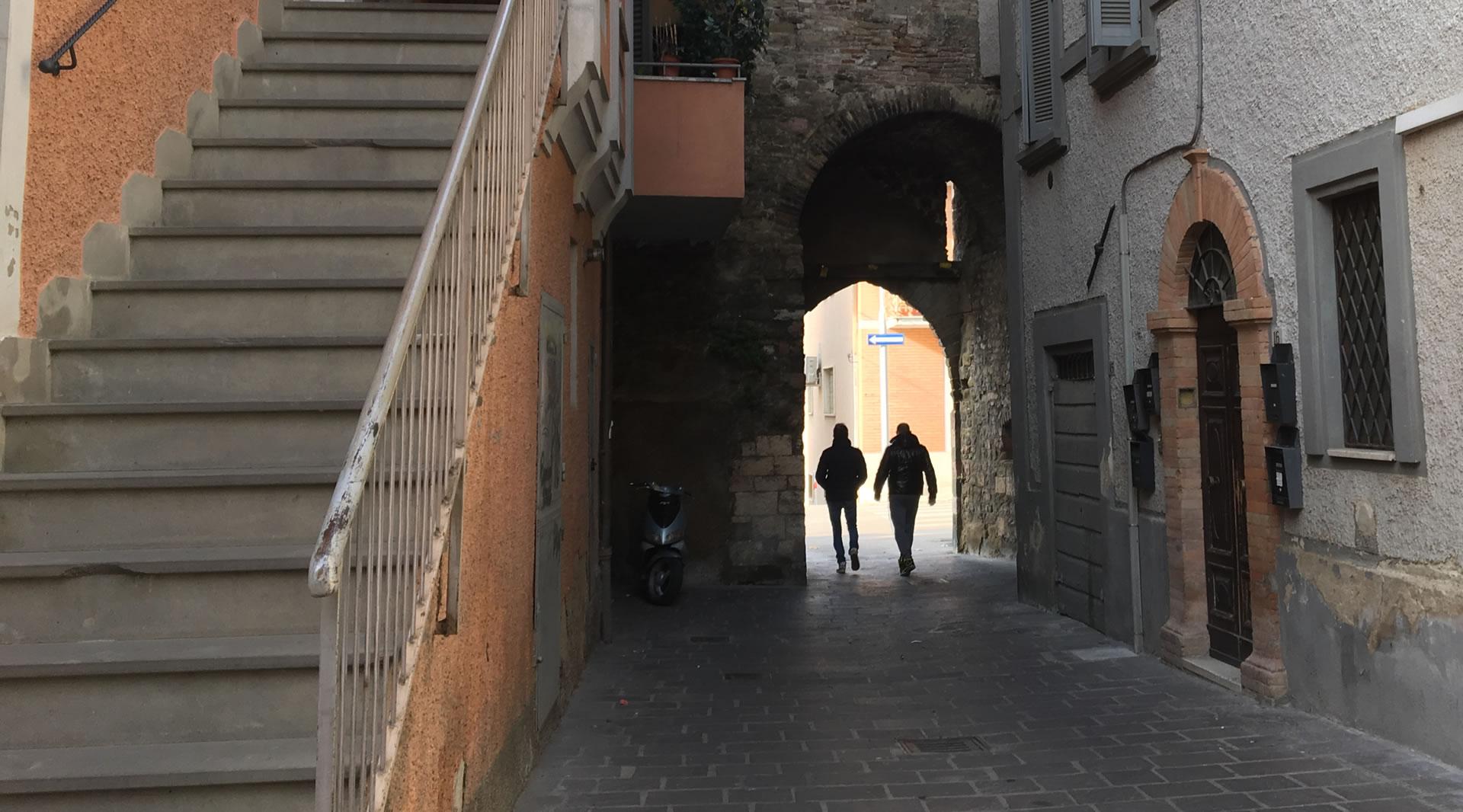 Due persone camminano sotto l’arco in pietra di Porta Sant’Angelo in un vicolo stretto e acciottolato, tra edifici storici con facciate in pietra e intonaco. Una scala esterna in pietra è visibile sulla sinistra.