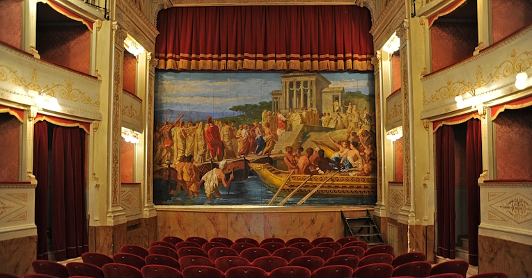 Interno del Teatro Clitunno di Trevi: poltrone rosse, palchetti decorati e sipario dipinto con una scena storica.