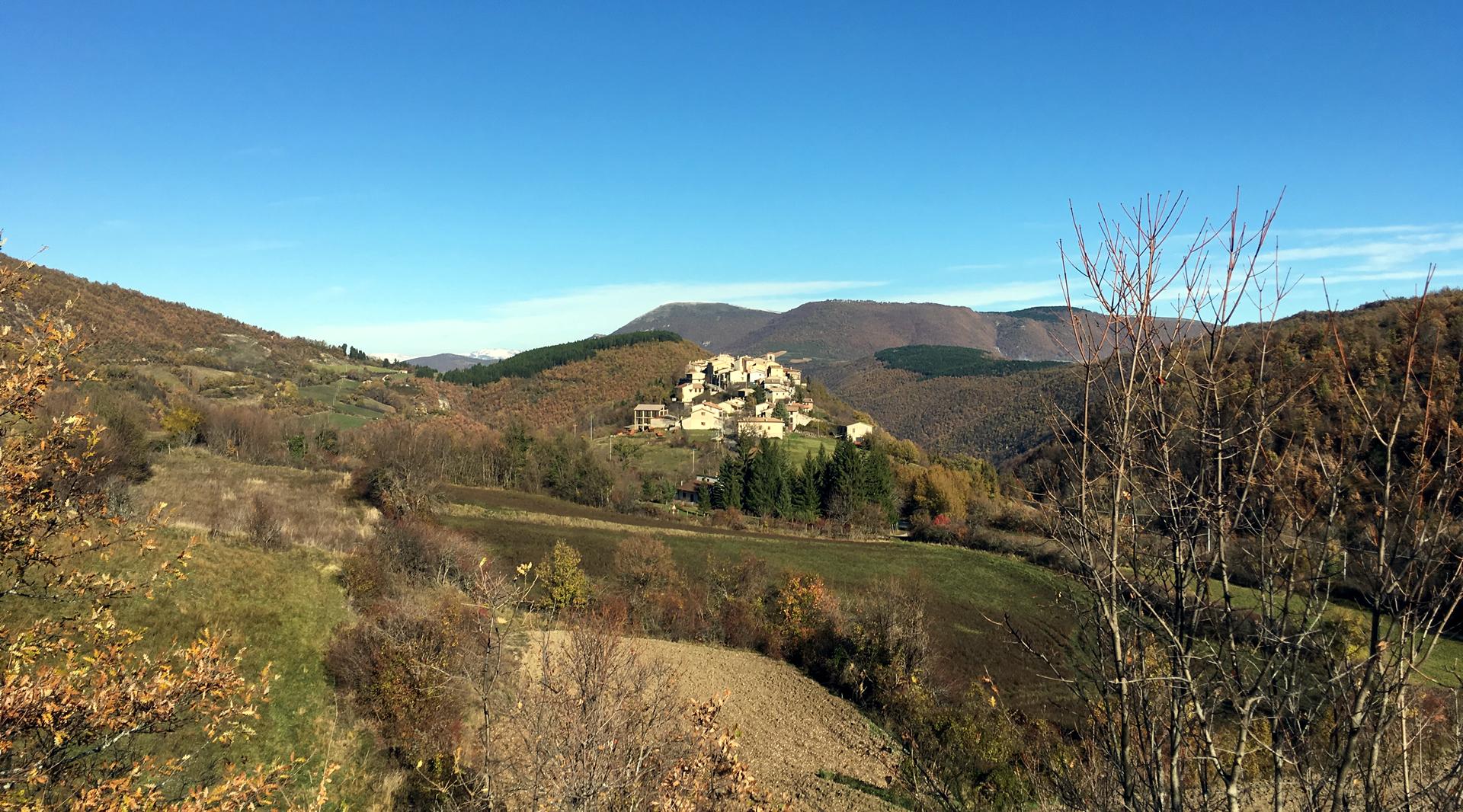 Blick auf die Burg von Usigni, die auf einem Hügel liegt, umgeben von grünen Bergen mit herbstlicher Vegetation und einem klaren Himmel.
