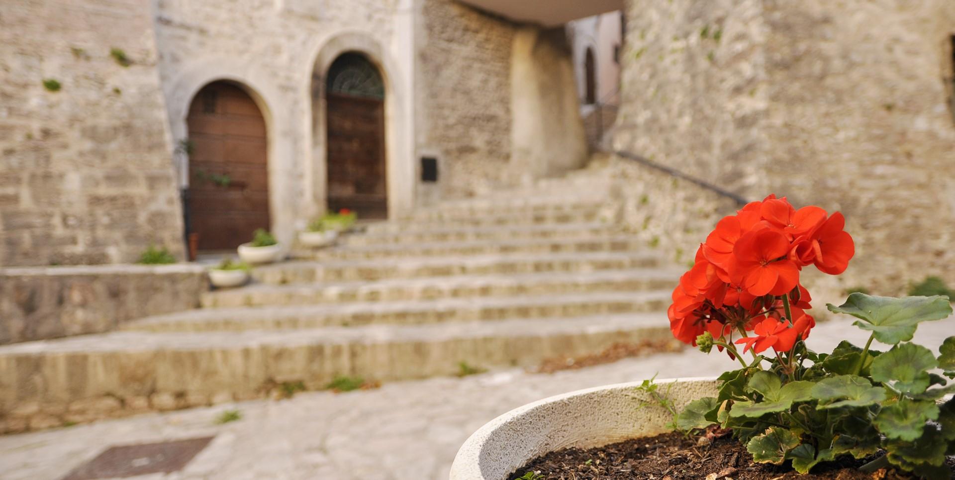 Eine steinerne Gasse mit Bögen, Treppen und historischen Häusern, mit einem Blumentopf mit roten Geranien im Vordergrund.