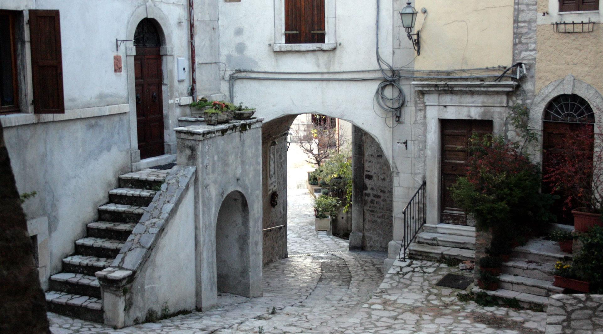 Eine steinerne Gasse mit Bögen, Treppen und historischen Häusern, die auf einen gepflasterten Platz führt.