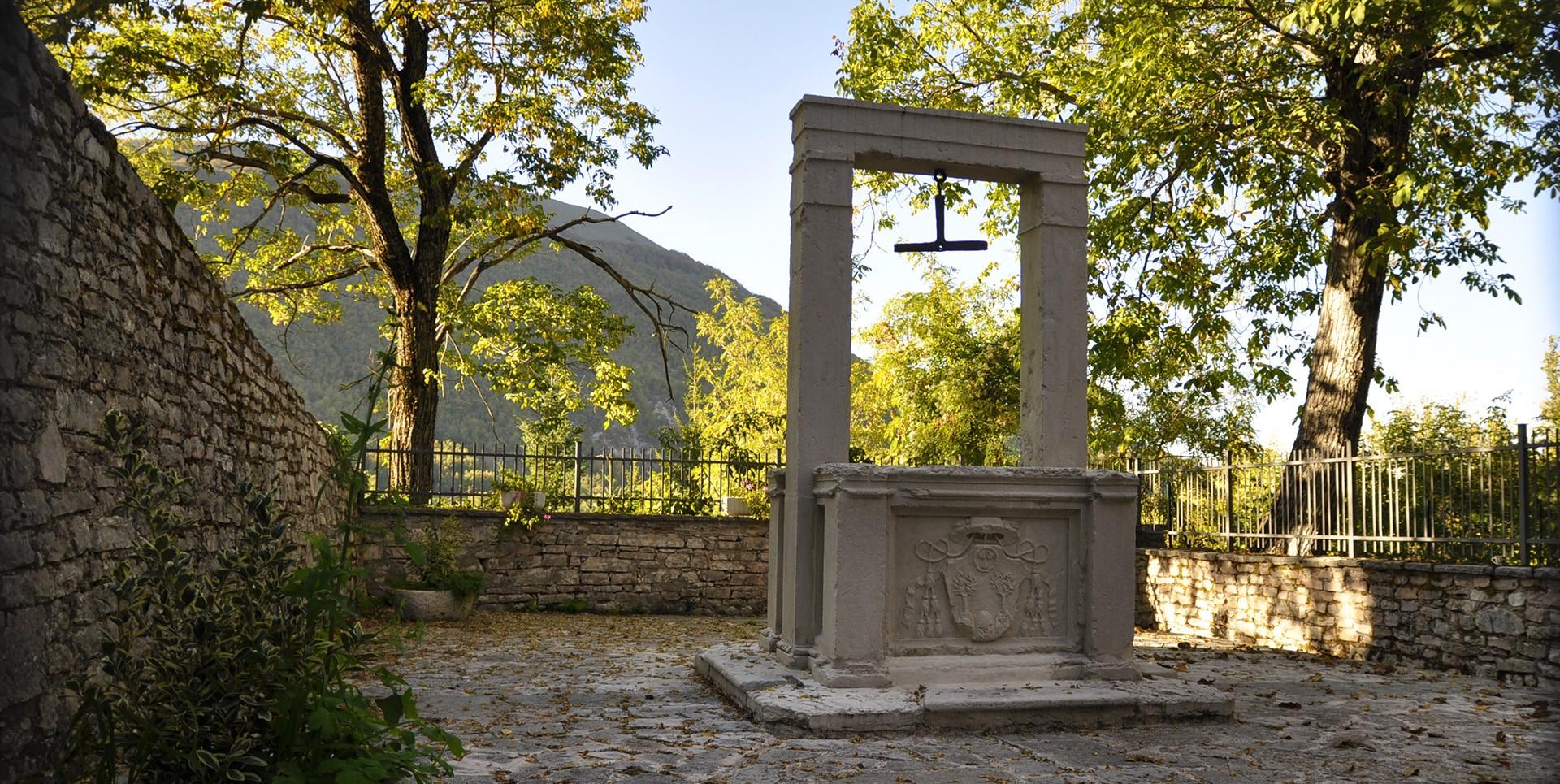 nullHistorischer Steinbrunnen mit Architrav und dem Adelswappen der Familie Poli, umgeben von Bäumen und Mauern in Usigni, Poggiodomo.