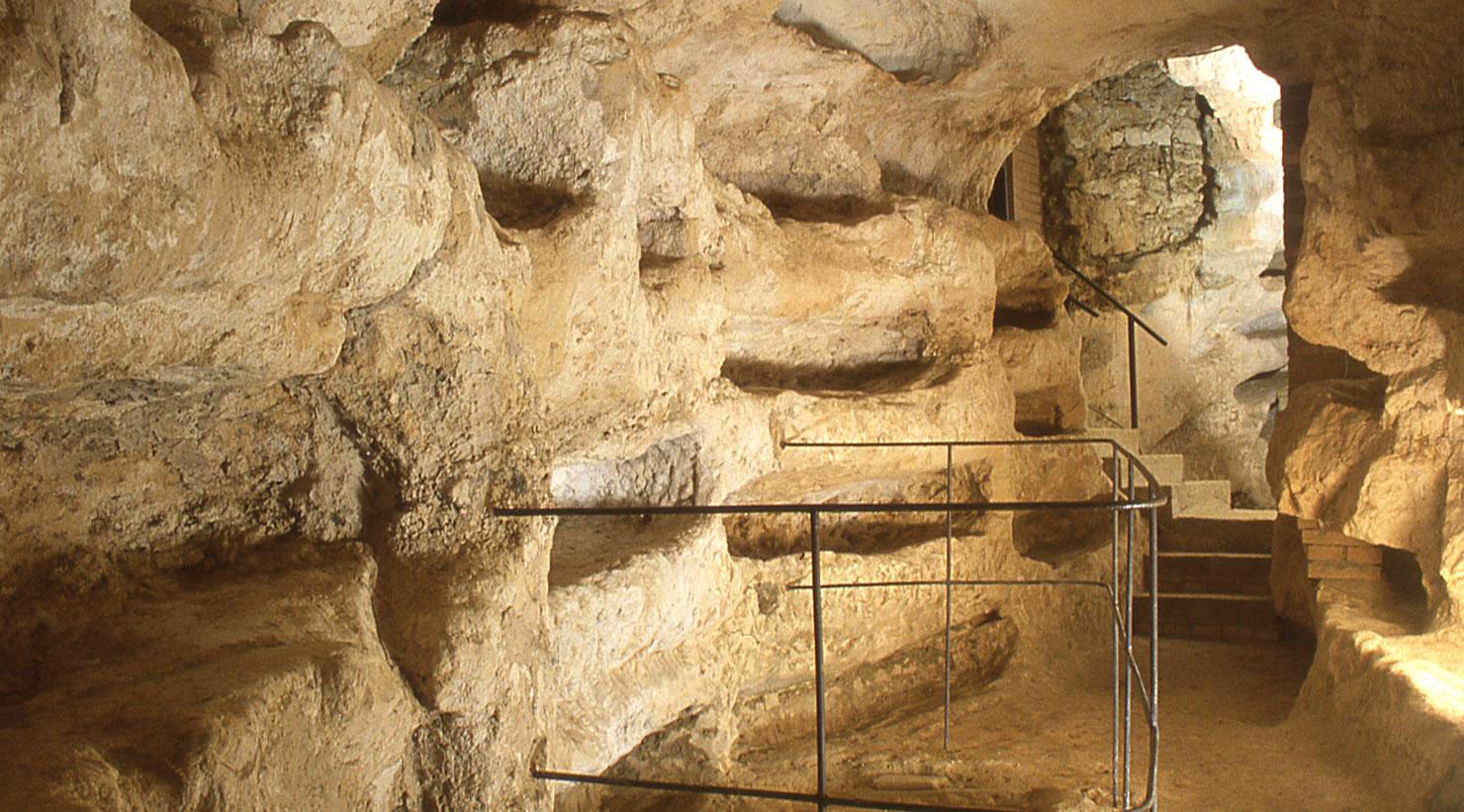 Entrance to an underground corridor of the Catacomb of Villa San Faustino. In the foreground, a narrow staircase with an iron handrail descends into a long corridor where burial niches and loculi can be observed carved into the brick and stone walls.