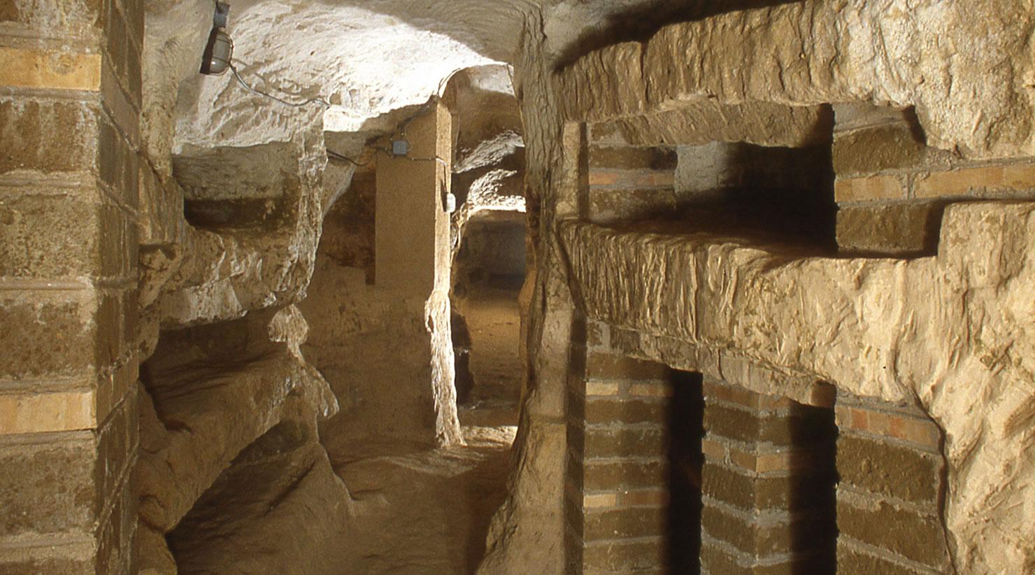 Interior of the Catacomb of Villa San Faustino. In the foreground, an underground corridor with natural rock walls where several burial niches of various sizes are carved. The corridor opens onto a staircase with an iron handrail leading to an upper level.