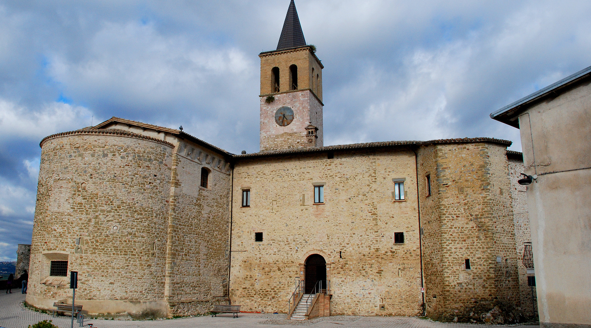 Stone church with apse, bell tower and clock, with a cloudy sky in the background