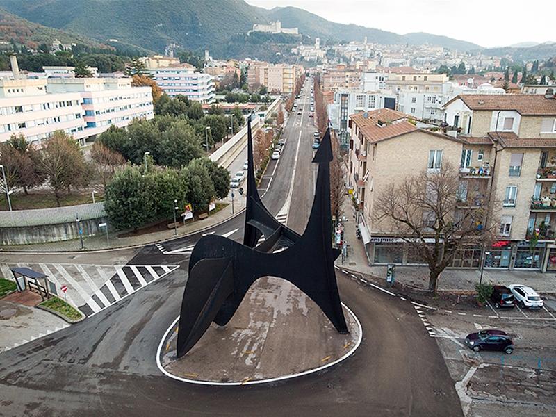 Luftaufnahme des Teodelapio von Alexander Calder im Kreisverkehr vor dem Bahnhof von Spoleto, mit der Allee und der Altstadt im Hintergrund.