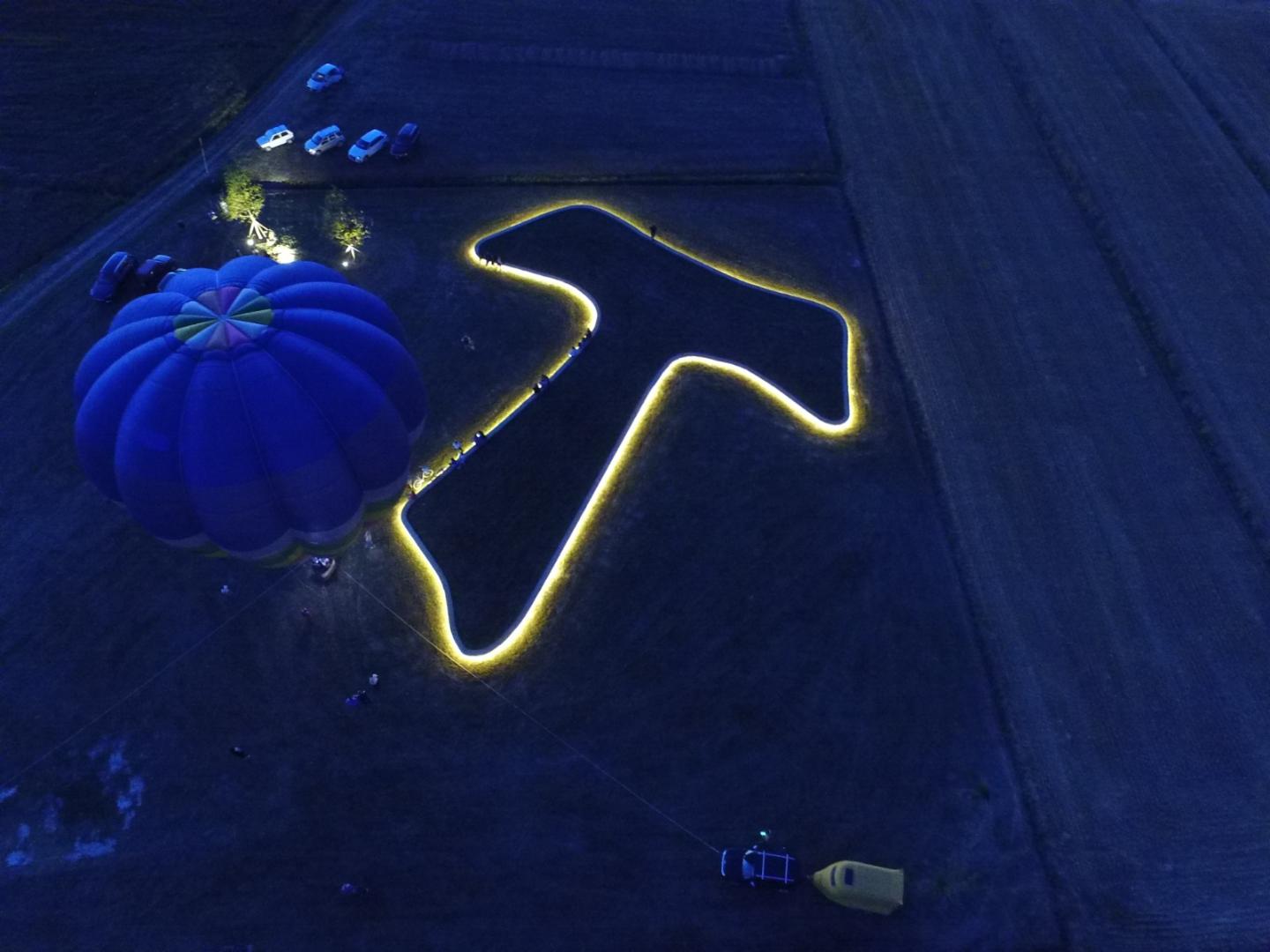 Vue nocturne du TAU de Piandarca illuminé, avec une montgolfière colorée et des voitures garées dans les champs alentours.