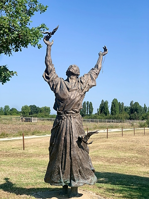 Statua di San Francesco con le braccia alzate e un uccello su ogni mano, in un campo soleggiato