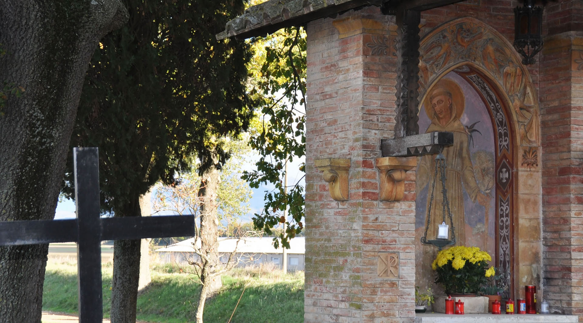 Edicola votiva con dipinto di san Francesco, fiori gialli e ceri rossi, tra alberi e vegetazione.