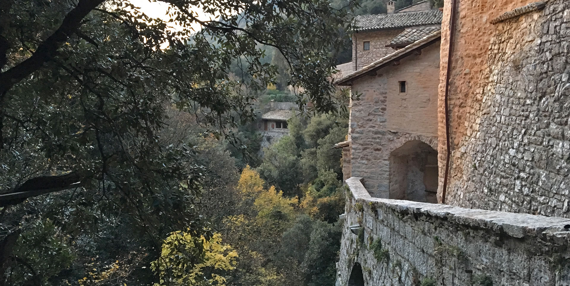 Vecchi muri in pietra affacciati su una valle verdeggiante con alberi e case nascoste tra le foglie autunnali