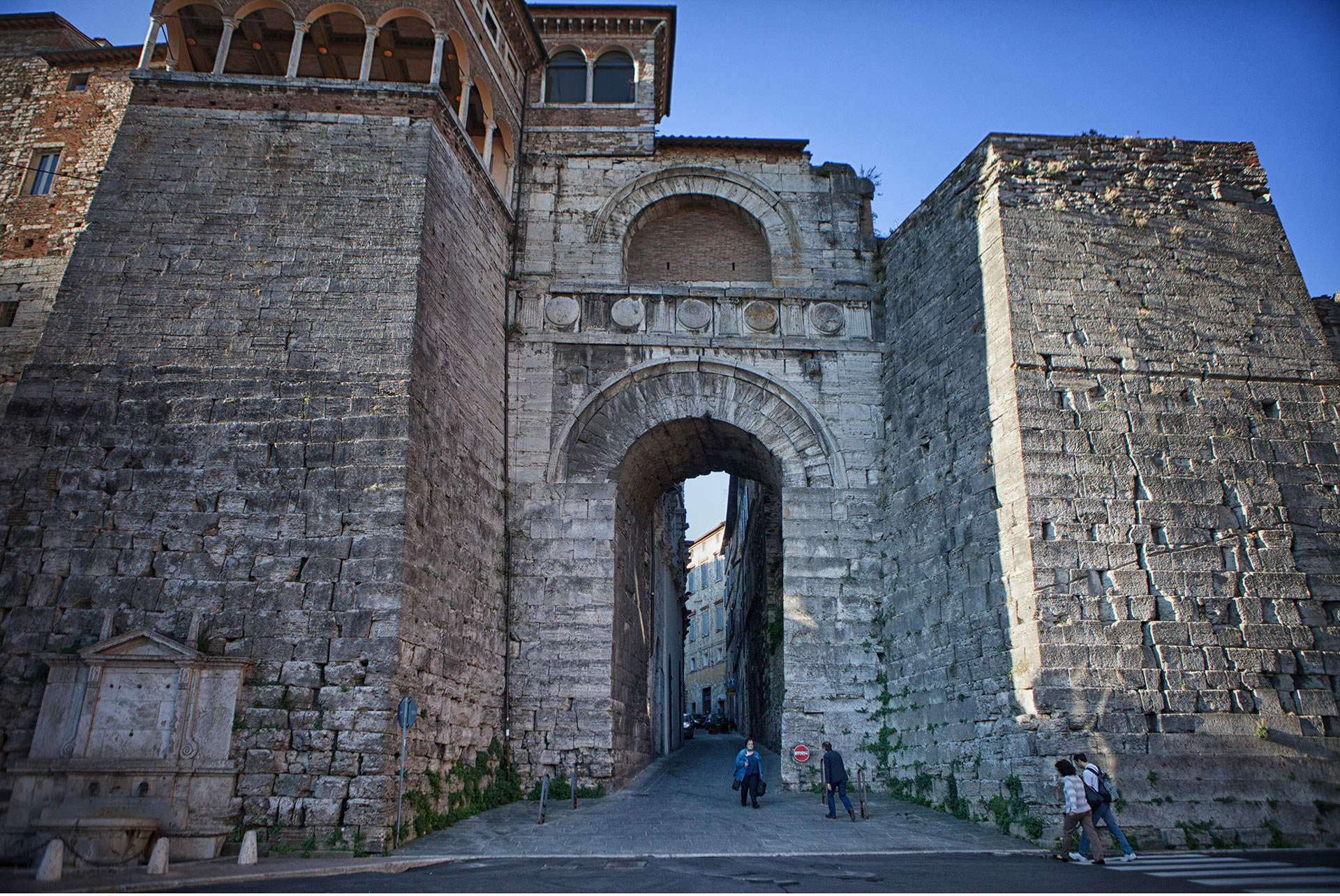 Arco etrusco in pietra con grande arco centrale, alte mura ai lati e persone che attraversano l’ingresso storico.