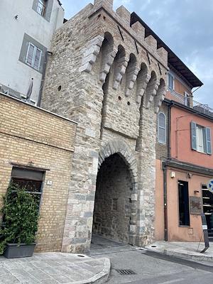 Antica Porta Sant'Angelo in pietra, con arco e merlature, incastonata tra edifici moderni a Bastia Umbra.
