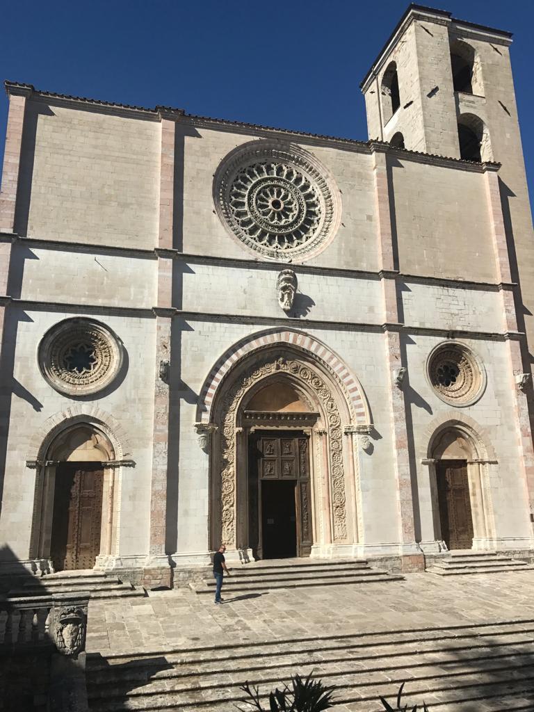 Façade of Todi Cathedral in light-coloured stone with a central rose window, decorated portal, and bell tower, under a clear sky.