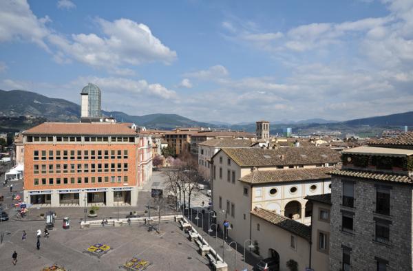  Panorama of Terni’s main square with modern and historic buildings, people walking, and Umbrian hills in the background. 