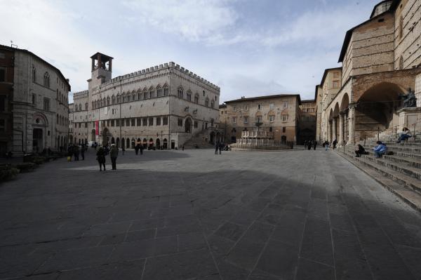  Historic square in Perugia with medieval buildings, a monumental fountain in the center, and people walking or sitting on the steps. 