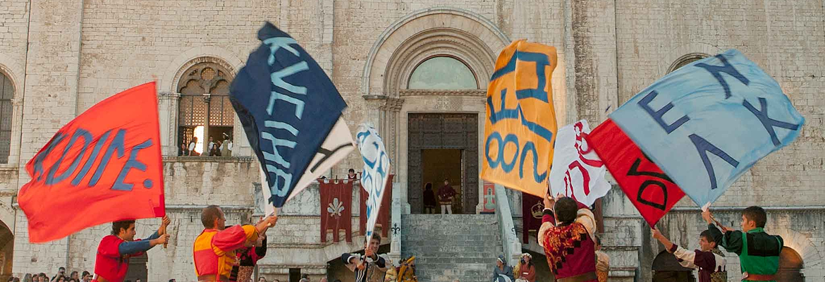 Fahnenschwinger in historischen Kostümen treten auf der Piazza Grande in Gubbio auf und schwenken bunte Fahnen vor einer Kirche.