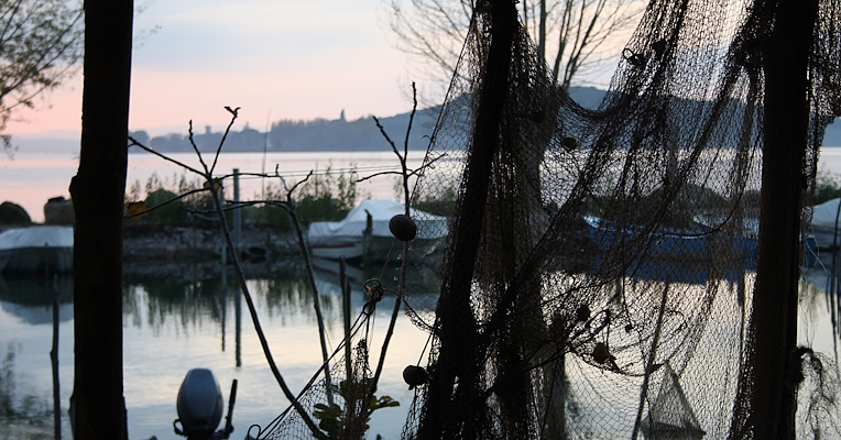 Reti da pesca appese incorniciano il lago Trasimeno al tramonto, con barche ormeggiate e un'isola sullo sfondo.