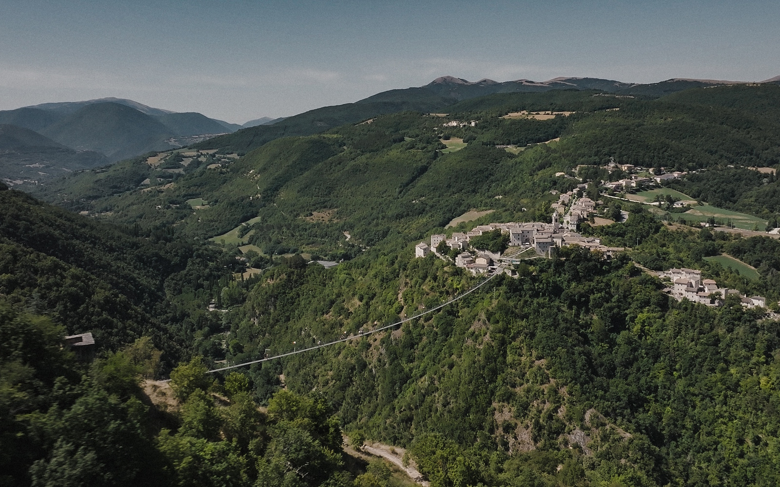 Veduta del ponte tibetano di Sellano sospeso tra due colline boscose, con il borgo arroccato e la vallata sullo sfondo.