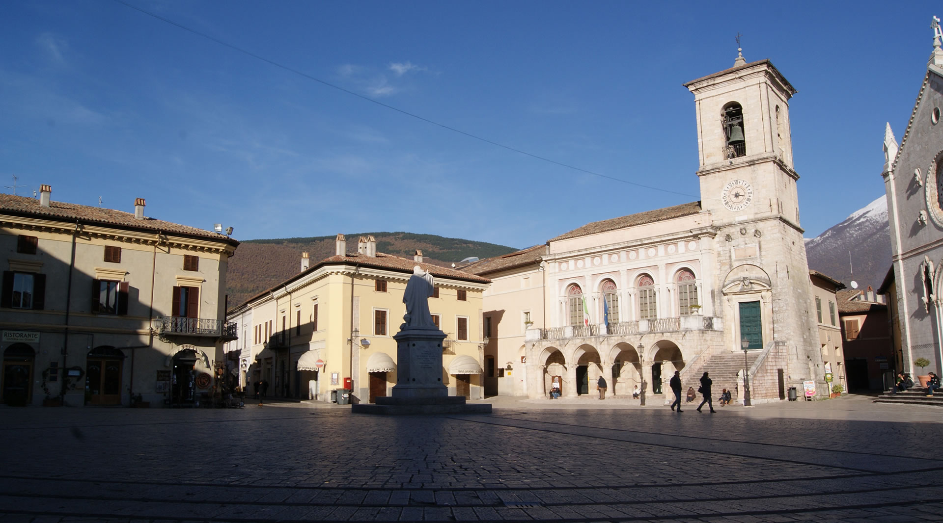Piazza San Benedetto in Norcia, with the statue of the saint, the Town Hall, and the bell tower, illuminated by sunlight.