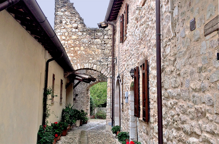 Stretta via medievale con archi e case in pietra, pavimentata in ciottoli e decorata con piante