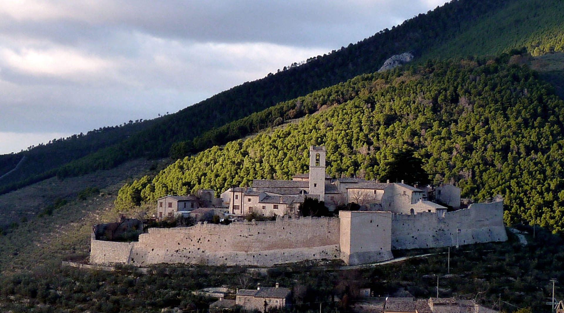Castello medievale di Campello Alto, con mura di cinta e campanile illuminato dal sole, circondato da montagne