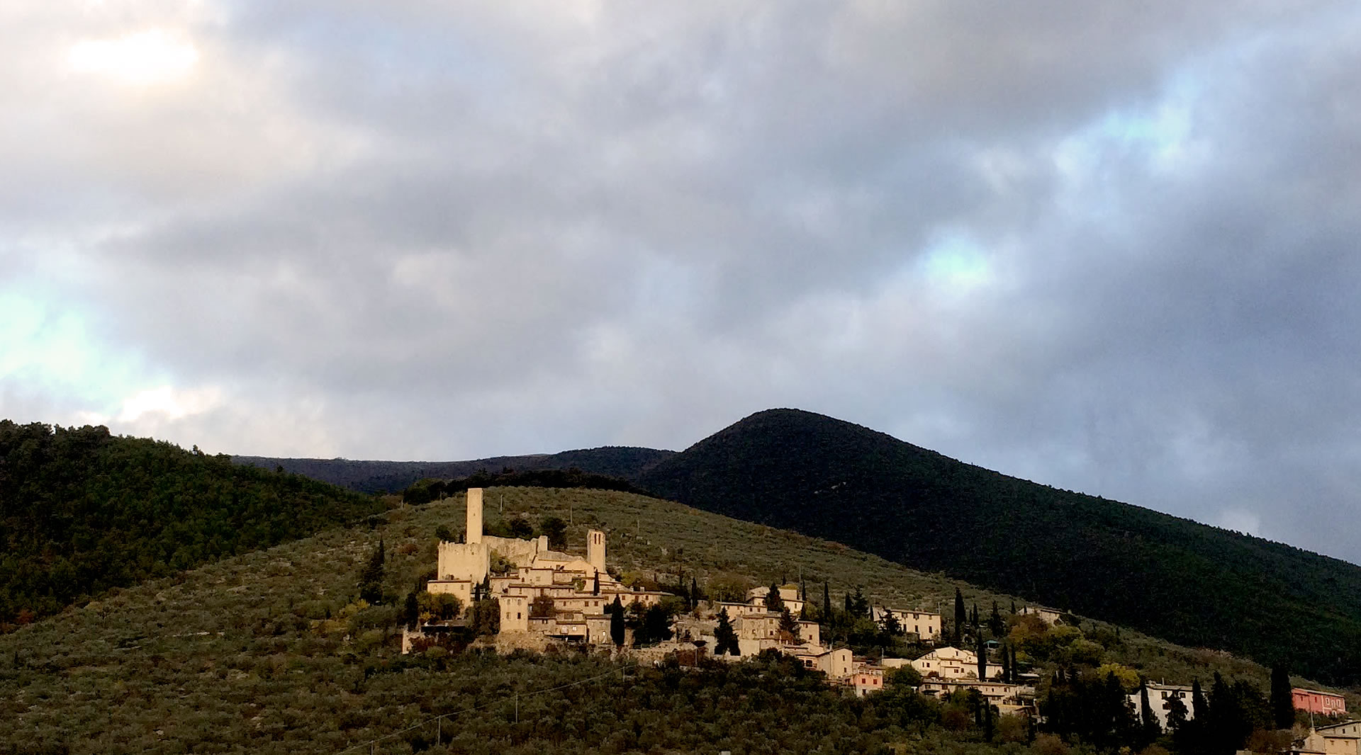 Antico castello medievale arroccato sulle colline, con torri e case in pietra, circondato da uliveti e montagne sotto un cielo nuvoloso