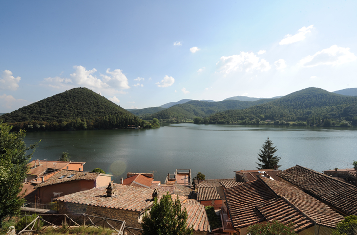 Blick auf den Lago di Piediluco mit Terrakottadächern im Vordergrund und grünen Hügeln, die sich im ruhigen Wasser spiegeln