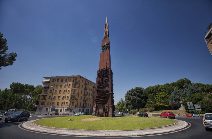 Der Obelisk Lancia di Luce im Kreisverkehr, mit Stadtverkehr und Wohngebäuden im Hintergrund