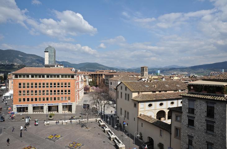 Panorama de la place principale de Terni avec des bâtiments modernes et historiques, des passants et les collines ombriennes en arrière-plan.