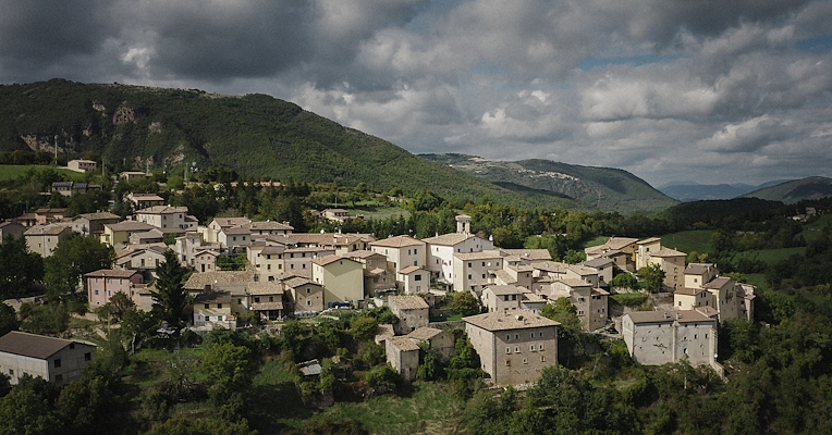 Vista aerea di Poggiodomo, con il centro storico circondato da colline verdi sotto un cielo nuvoloso.
