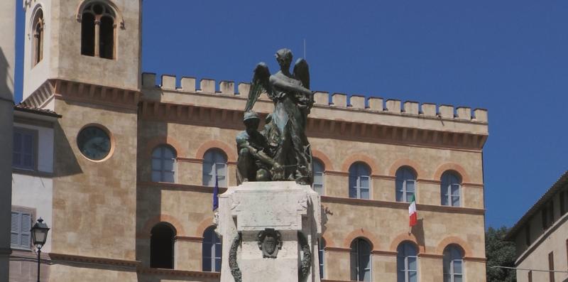 Statua commemorativa in bronzo con un soldato e un angelo al centro di una piazza, con un edificio storico sullo sfondo.