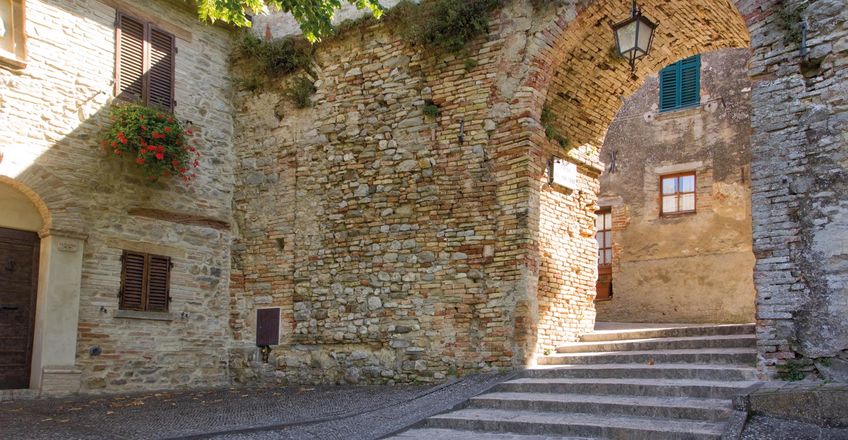 Mittelalterlicher Steinbogen in Montone, mit sonnenbeschienener Treppe und historischen Häusern mit Fenstern und Läden.