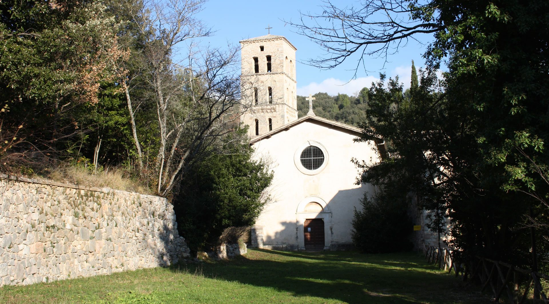 Abbey surrounded by greenery, featuring a simple facade and a Romanesque stone bell tower, enclosed by trees and stone walls.