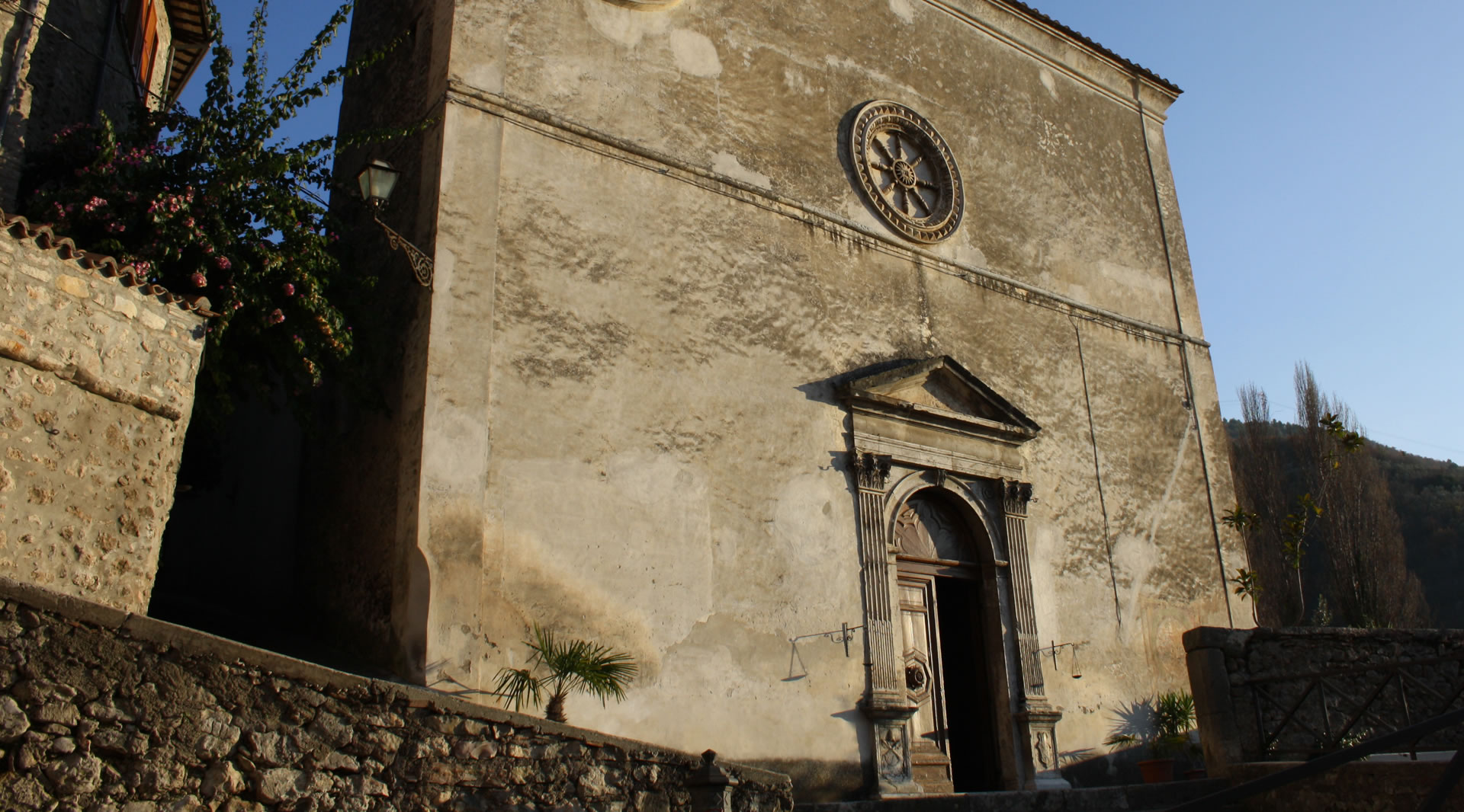 Ancient church with a weathered facade, stone portal, and rose window, set among stone walls and lush vegetation.