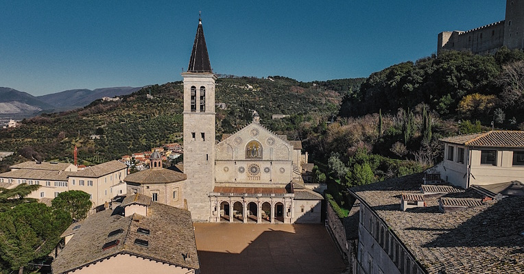 Luftaufnahme der Kathedrale von Spoleto mit romanischer Fassade, Glockenturm und Platz, umgeben von Hügeln und historischen Dächern.
