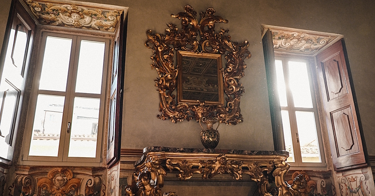 Interno barocco con specchiera dorata, consolle scolpita e affreschi decorativi tra due finestre luminose.