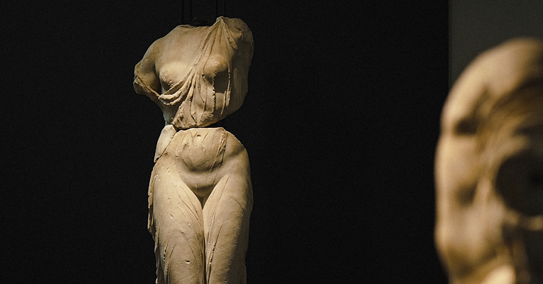 Scultura classica femminile priva di testa e braccia, esposta in un museo su sfondo scuro.