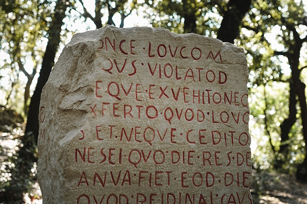 Epigrafe latina incisa su pietra antica nel bosco di Monteluco, con lettere rosse ben visibili tra gli alberi.