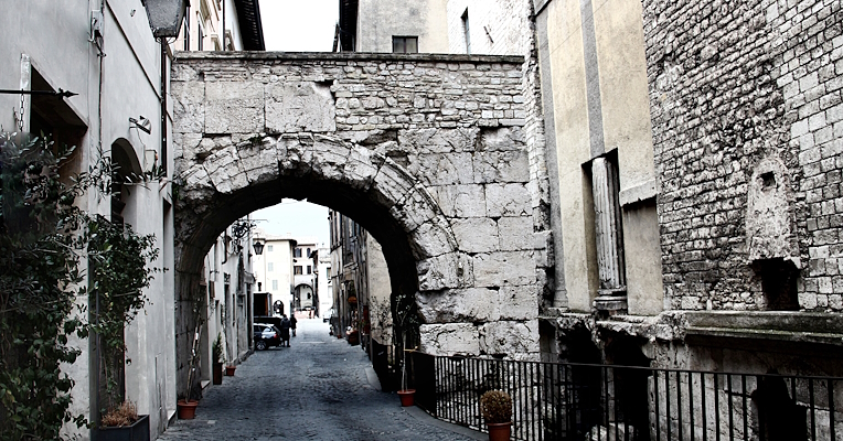 Arco romano in pietra in una stretta via di Spoleto, con edifici storici ai lati che conservano resti di colonne romane