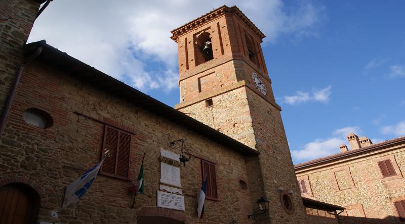 Palazzo del Municipio di Paciano in mattoni, con torre dell'orologio e campanile, sotto un cielo parzialmente nuvoloso.