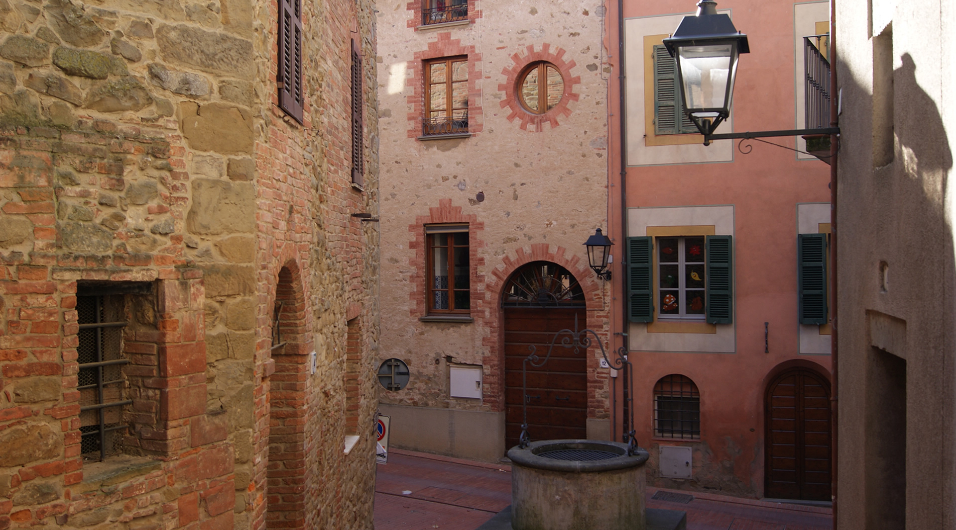 Narrow street in the medieval village of Paciano with stone and brick buildings. In the center, a circular stone well.