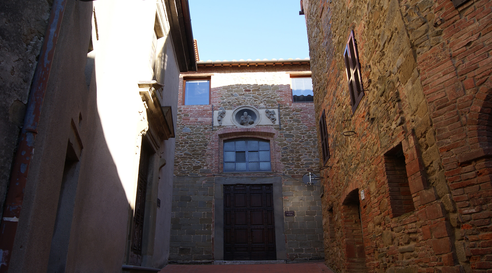 Stone facade of a church located in a medieval street, with a central door and decoration above the entrance.